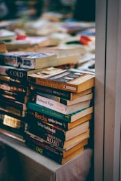 A large stack of paperback books is neatly arranged on a table and appears to be part of a larger book display or sale. The spines of the books show a variety of colors and fonts, with some titles and authors clearly visible. The background contains more blurred stacks of books, suggesting a busy and crowded atmosphere.