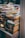 A large stack of paperback books is neatly arranged on a table and appears to be part of a larger book display or sale. The spines of the books show a variety of colors and fonts, with some titles and authors clearly visible. The background contains more blurred stacks of books, suggesting a busy and crowded atmosphere.