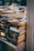 A large stack of paperback books is neatly arranged on a table and appears to be part of a larger book display or sale. The spines of the books show a variety of colors and fonts, with some titles and authors clearly visible. The background contains more blurred stacks of books, suggesting a busy and crowded atmosphere.