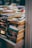 A large stack of paperback books is neatly arranged on a table and appears to be part of a larger book display or sale. The spines of the books show a variety of colors and fonts, with some titles and authors clearly visible. The background contains more blurred stacks of books, suggesting a busy and crowded atmosphere.
