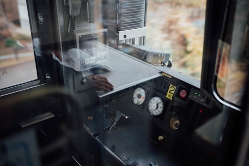 A train control cabin featuring a dashboard with dials, buttons, and communication equipment. Reflections are visible on the glass, and a person's hand is seen on the surface of the panel.