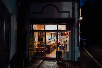 A cozy, warmly lit exterior of a small restaurant at night, showcasing a welcoming interior with wooden chairs and tables neatly arranged. The entrance is adorned with various signs, and the soft glow from inside contrasts with the dark street outside.