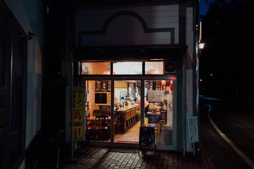 A cozy, warmly lit exterior of a small restaurant at night, showcasing a welcoming interior with wooden chairs and tables neatly arranged. The entrance is adorned with various signs, and the soft glow from inside contrasts with the dark street outside.
