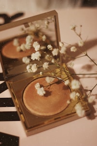 A compact makeup product with a golden case lies open on a surface, accompanied by delicate white flowers scattered around and reflected in the mirror above the makeup. The surface has a soft, warm tone, complementing the overall aesthetic.