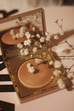 A compact makeup product with a golden case lies open on a surface, accompanied by delicate white flowers scattered around and reflected in the mirror above the makeup. The surface has a soft, warm tone, complementing the overall aesthetic.