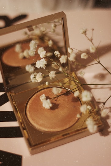 A compact makeup product with a golden case lies open on a surface, accompanied by delicate white flowers scattered around and reflected in the mirror above the makeup. The surface has a soft, warm tone, complementing the overall aesthetic.