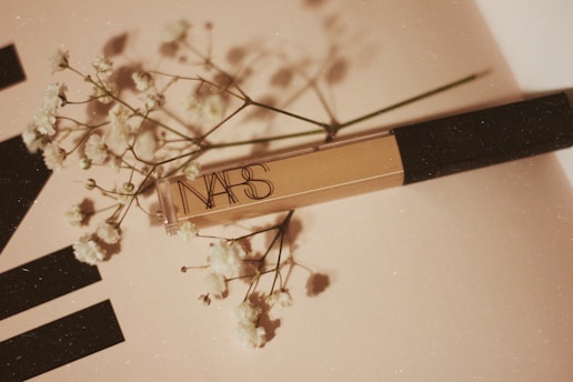 A NARS cosmetic product placed on a light-colored surface accompanied by delicate white flowers, creating a soft and elegant composition.
