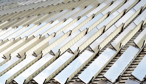 Reflective metal panels are arranged in a repeating diagonal pattern, creating a striking geometric design. The panels reflect the sky, adding an element of natural light and depth to the image. The lines and angles form a modern architectural aesthetic.