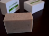A rectangular bar of soap with a textured surface sits on a dark wooden surface. The soap is beige with small specks, suggesting natural ingredients. In the background, two white boxes with green labels are slightly out of focus.