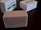 A rectangular bar of soap with a textured surface sits on a dark wooden surface. The soap is beige with small specks, suggesting natural ingredients. In the background, two white boxes with green labels are slightly out of focus.
