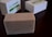 A rectangular bar of soap with a textured surface sits on a dark wooden surface. The soap is beige with small specks, suggesting natural ingredients. In the background, two white boxes with green labels are slightly out of focus.