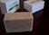 A rectangular bar of soap with a textured surface sits on a dark wooden surface. The soap is beige with small specks, suggesting natural ingredients. In the background, two white boxes with green labels are slightly out of focus.