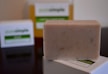 A bar of natural, handcrafted soap is placed in the foreground, with its texture showing small flecks of ingredients. In the background, two soap boxes are slightly blurred, with green and white labels, indicating a focus on natural or organic ingredients.