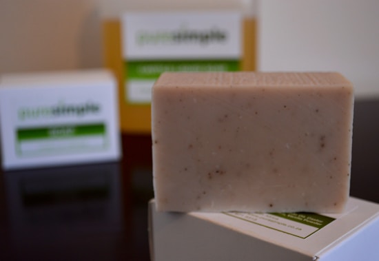 A bar of natural, handcrafted soap is placed in the foreground, with its texture showing small flecks of ingredients. In the background, two soap boxes are slightly blurred, with green and white labels, indicating a focus on natural or organic ingredients.