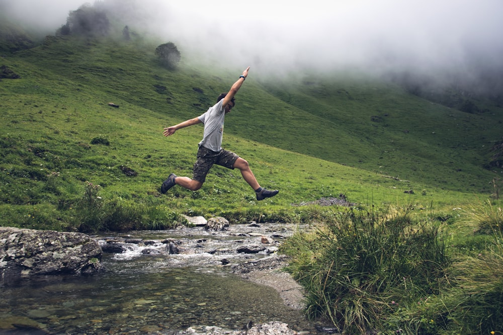 man jumping above body of water between green grass during daytime