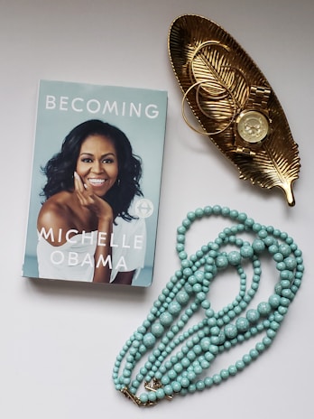 A book titled 'Becoming' with a portrait of a smiling woman on the cover is placed next to a golden leaf-shaped tray. The tray holds a round golden watch and two bangles. Below them, there is a necklace made of pale turquoise beads.