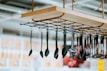 A range of kitchen utensils, including ladles and spatulas, hang from a suspended wooden rack with a metal grid and rope supports. The background is slightly blurred, indicating an indoor setting with various displays.