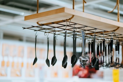 A range of kitchen utensils, including ladles and spatulas, hang from a suspended wooden rack with a metal grid and rope supports. The background is slightly blurred, indicating an indoor setting with various displays.