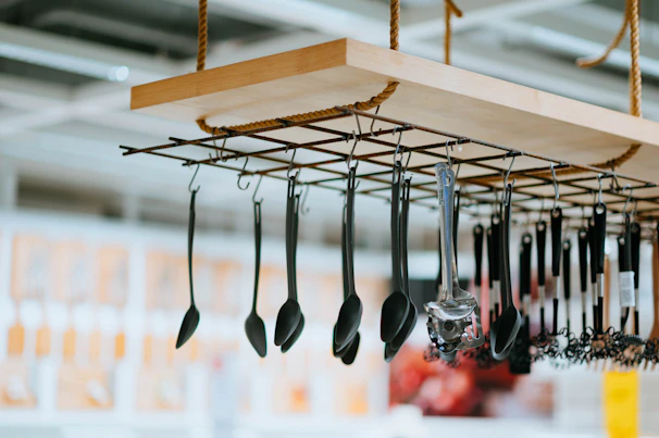 A range of kitchen utensils, including ladles and spatulas, hang from a suspended wooden rack with a metal grid and rope supports. The background is slightly blurred, indicating an indoor setting with various displays.