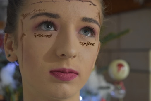 A person with artistic writing and designs on their face, looking slightly upwards with a thoughtful expression. The background features a blurred setting, possibly indicating a holiday or festive environment.