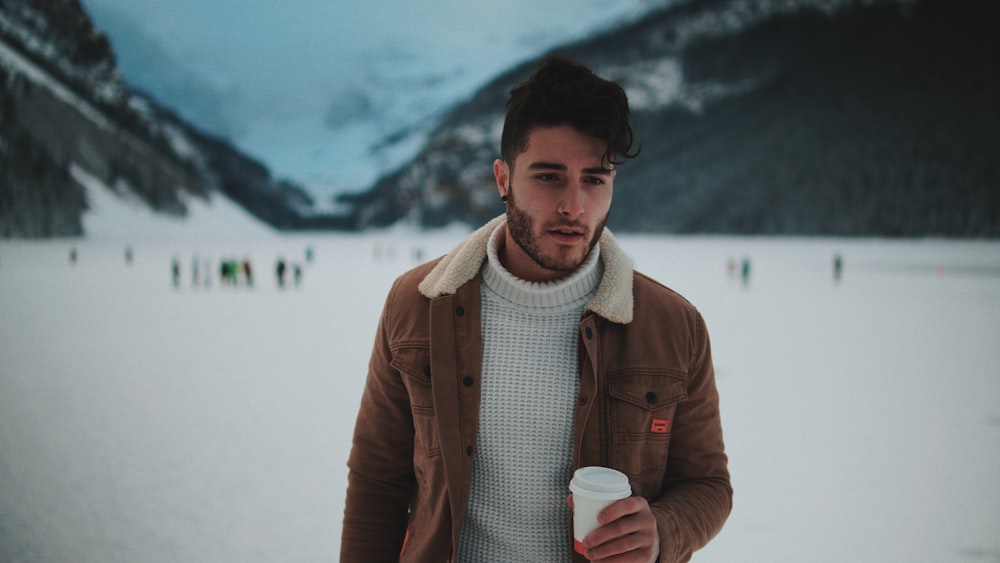 man in brown jacket holds cup with lid near people standing on snow covered ground