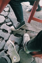 Two pairs of casual sneakers on hexagonal stone pavement, with the lower half of people's legs visible. One person wears black sneakers, and the other wears gray sneakers. A red chair is partially visible, and the scene has an outdoor, casual vibe.