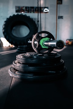 A dimly lit gym setting featuring stacked weight plates and a barbell in the foreground. A large tire is visible in the background, along with some gym equipment. The atmosphere is industrial, with shadows creating a moody ambiance.