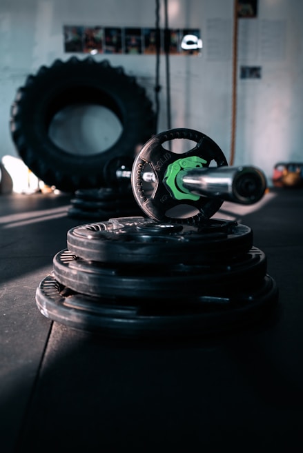 A dimly lit gym setting featuring stacked weight plates and a barbell in the foreground. A large tire is visible in the background, along with some gym equipment. The atmosphere is industrial, with shadows creating a moody ambiance.