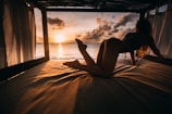 A person is posed on a bed in a cabana, with an ocean sunset backdrop. The sky displays warm hues as the sun sets on the horizon, casting shadows across the scene. The ambiance is relaxed and serene, with curtains framing the open view of the sea.