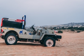A vintage open-top jeep is parked on desert sand, with a backdrop of arid landscape and distant hills. The jeep has a sign on the side indicating 'Jeep Safari' with blue camel graphics.