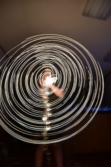 Concentric circles of bright light are spiraling outward, creating a dynamic and mesmerizing pattern against a dark background. A blurred figure can be partially seen through the glowing swirls, suggesting movement and energy.