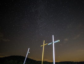 Three crosses stand against a dark, starry night sky. The scene is illuminated from below, casting light on the crosses, while a silhouette of a forested hill is visible in the background.