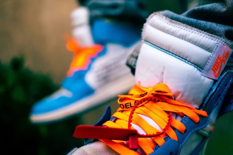 A close-up view of a pair of sneakers with vibrant orange laces, a blue and white color scheme, and a visible 'Nike Air' logo. One shoe has a red zip tie attached, and the sneakers appear to be worn by a person wearing jeans.