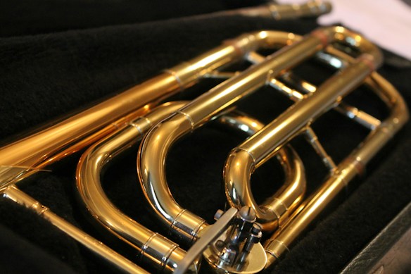 A close-up view of a brass musical instrument, focusing on its coiled tubing and shiny metal surface. The instrument is placed on a dark, possibly velvet, background that contrasts with its polished golden color.