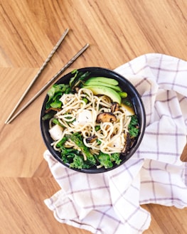 A bowl of noodles with mushrooms, tofu, and green vegetables, served on a black dish. The meal is on top of a beige checkered cloth and placed on a light wooden surface. Metal chopsticks are positioned next to the bowl.