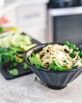 A black bowl filled with noodles, diced tofu, and a variety of green vegetables such as spinach and broccoli sits on a marble countertop. Fresh herbs and sliced vegetables are visible on a blurred cutting board in the background.