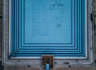 Aerial view of a large, empty swimming pool with blue tiled patterns creating a geometric design. The pool appears unused, with no water visible and some dirt or stains marked across its surface. Surrounding the pool edge are concrete walkways.