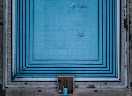 Aerial view of a large, empty swimming pool with blue tiled patterns creating a geometric design. The pool appears unused, with no water visible and some dirt or stains marked across its surface. Surrounding the pool edge are concrete walkways.