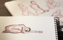 A sketchbook displaying a figure drawing of a person in a curled, reclining position. Surrounding pages show additional figure sketches. The artwork is rendered in reddish-brown pencil or crayon, with subtle shading capturing the human form.