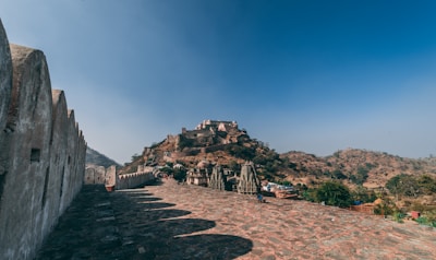 A majestic fort atop a hill surrounded by lush greenery and rocky terrain under a clear blue sky. The foreground features an ancient stone wall running parallel to a pathway leading toward the fort, with detailed stonework and battlements.