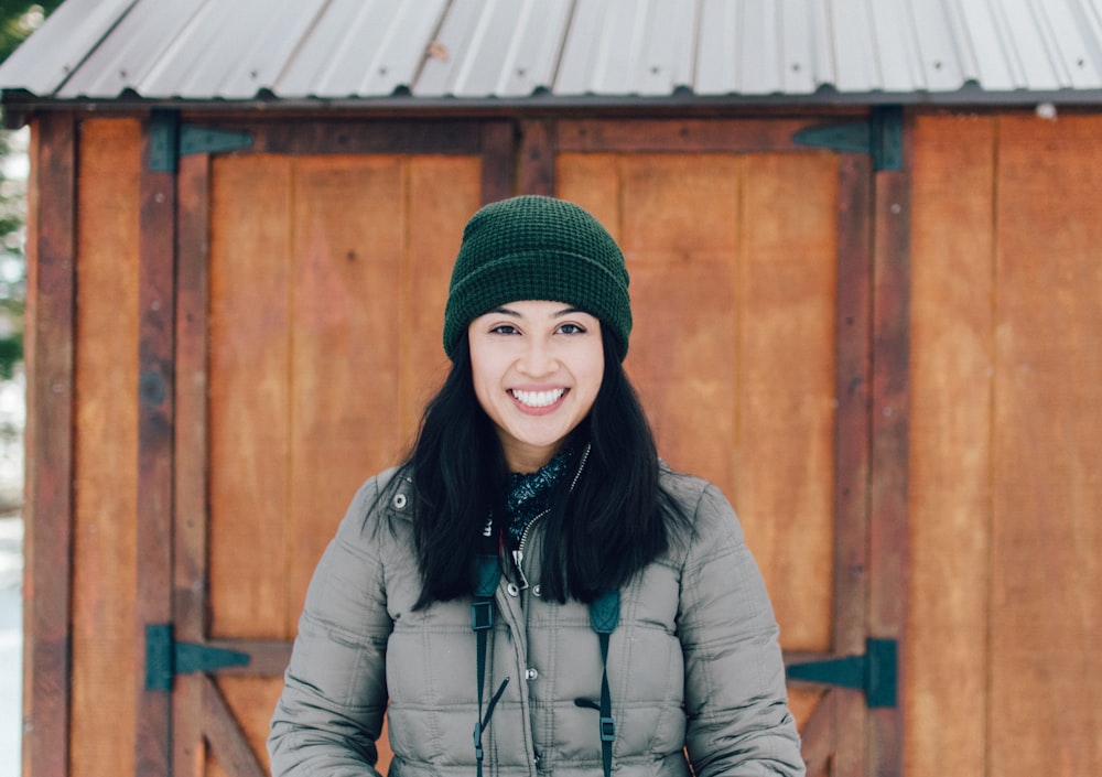 Mujer sonriente con chaqueta marrón de pie junto al cobertizo de madera marrón