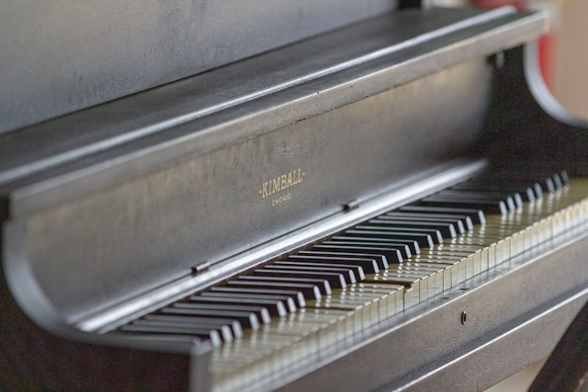 A close-up view of a dark colored piano with visible keys. The manufacturer's name, Kimball, is embossed on the piano. The focus highlights the sleek and polished finish of the instrument.
