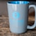 A gray mug with a blue interior is placed on a rustic wooden surface. The mug features a blue logo with a microphone icon and the words 'Ear Blowing Audio Productions' printed in bold letters. The logo stands out against the smooth, glossy finish of the mug.