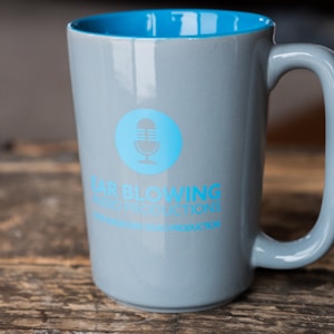 A gray mug with a blue interior is placed on a rustic wooden surface. The mug features a blue logo with a microphone icon and the words 'Ear Blowing Audio Productions' printed in bold letters. The logo stands out against the smooth, glossy finish of the mug.
