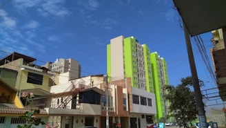A cityscape featuring mid-rise residential buildings with a modern design, colored in beige and bright green tones. The foreground includes smaller residential buildings with slanted roofs and external staircases, set against a clear blue sky.