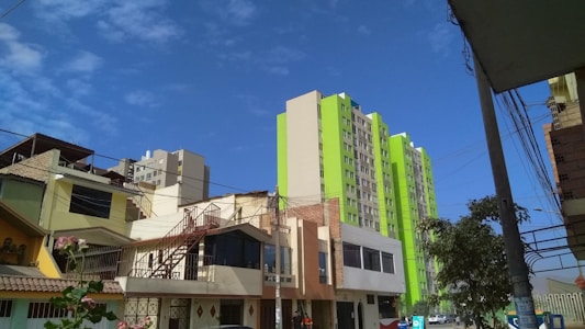 A cityscape featuring mid-rise residential buildings with a modern design, colored in beige and bright green tones. The foreground includes smaller residential buildings with slanted roofs and external staircases, set against a clear blue sky.