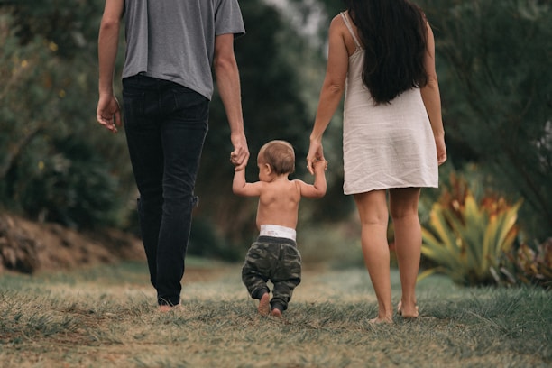 A toddler walks outdoors on a grassy path, holding hands with two adults, one on each side. The setting is lush and natural, with lots of greenery and plants visible in the background. The toddler appears to be learning to walk, supported by the adults.