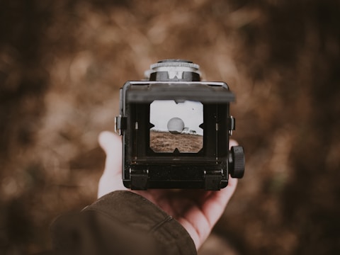 A hand holding a vintage camera with a viewfinder displaying an image of a landscape that includes a large, white spherical object and some structures. The background is blurred, making the focus on the camera and the viewfinder.