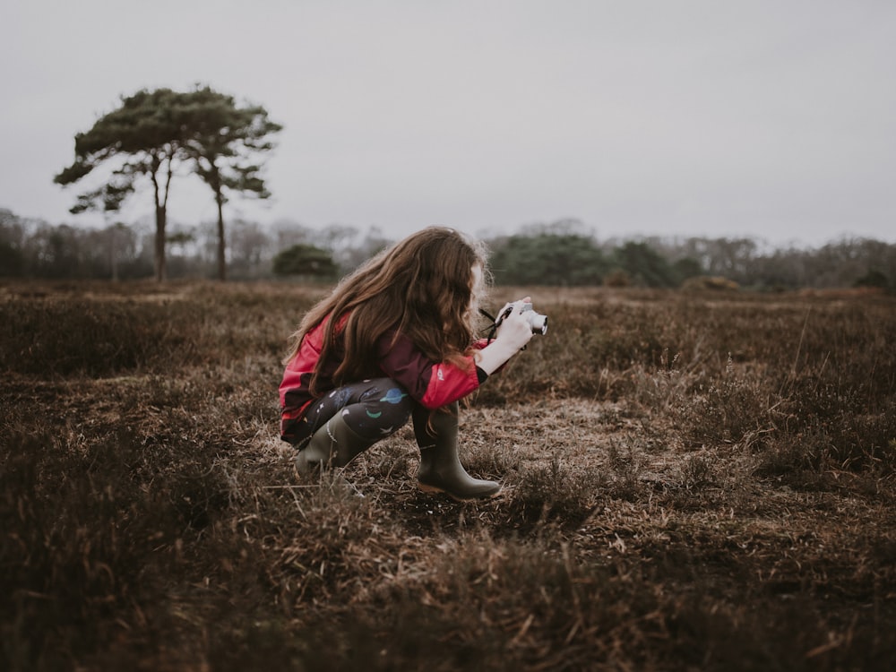 girl in red long-sleeved shirt sitting on stone while capturing grass field