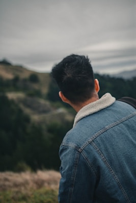 A person wearing a denim jacket with a fluffy collar stands outdoors, looking towards a scenic landscape with rolling hills and a cloudy sky. The scene appears calm and natural.